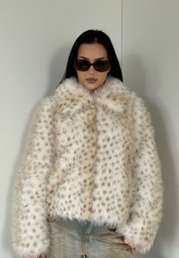 Veste en fausse fourrure avec une base beige et des taches foncées, dotée d'un col montant et de manches larges, associée à des lunettes de soleil foncées.