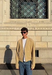 Črtast rumen in bel pulover na gumb, širokih rokavov. Kombiniran z Belo srajco in modrimi kavbojkami, postavljen pred teksturirano steno.