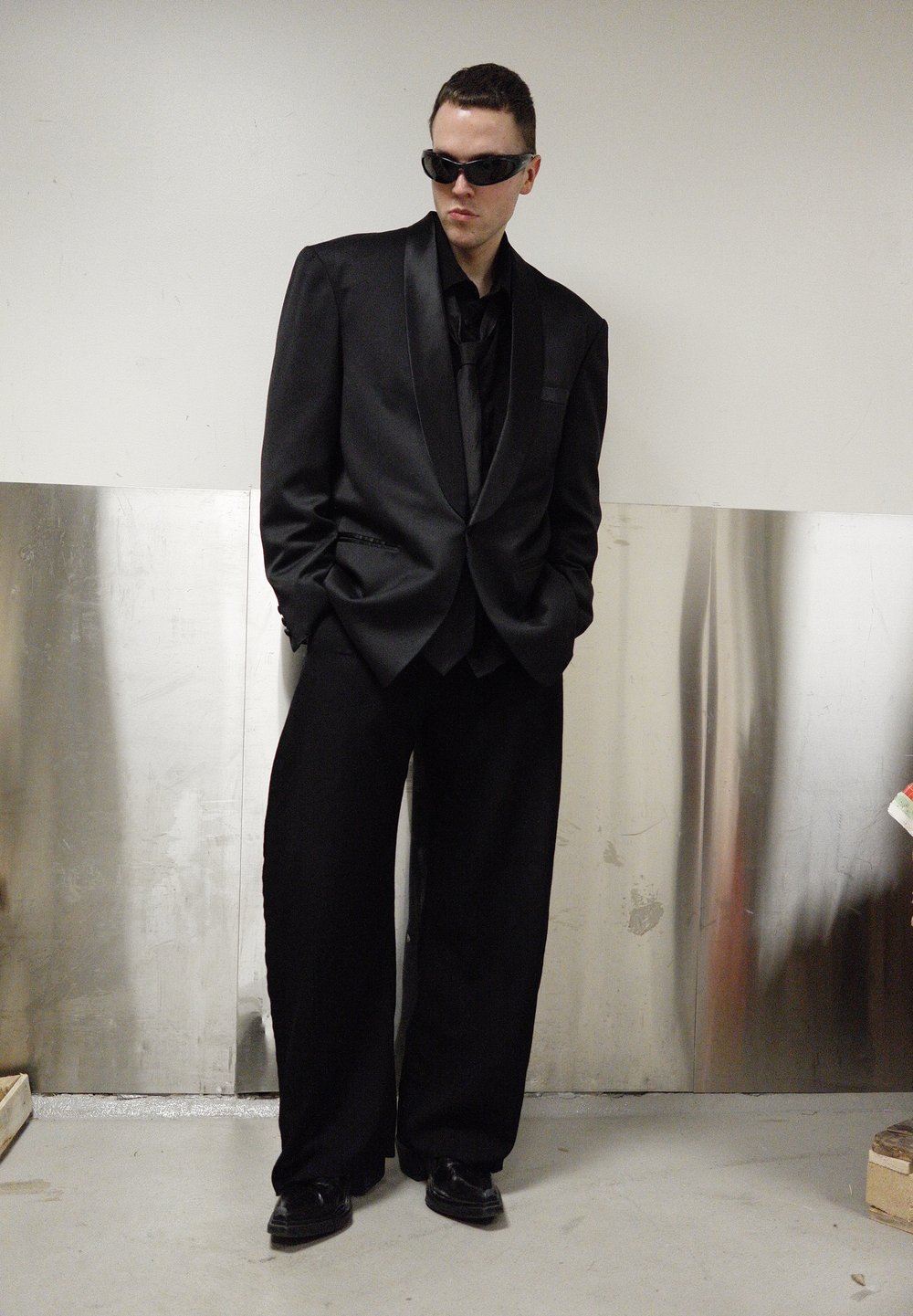 Costume noir oversize avec un revers en satin, accompagné d'un pantalon noir ample. Les accessoires comprennent des lunettes de soleil noires et des chaussures polies.