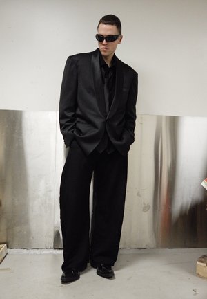 Traje negro de gran tamaño con solapa de satén, combinado con pantalones negros de corte holgado. Los accesorios incluyen gafas de sol negras y zapatos pulidos.