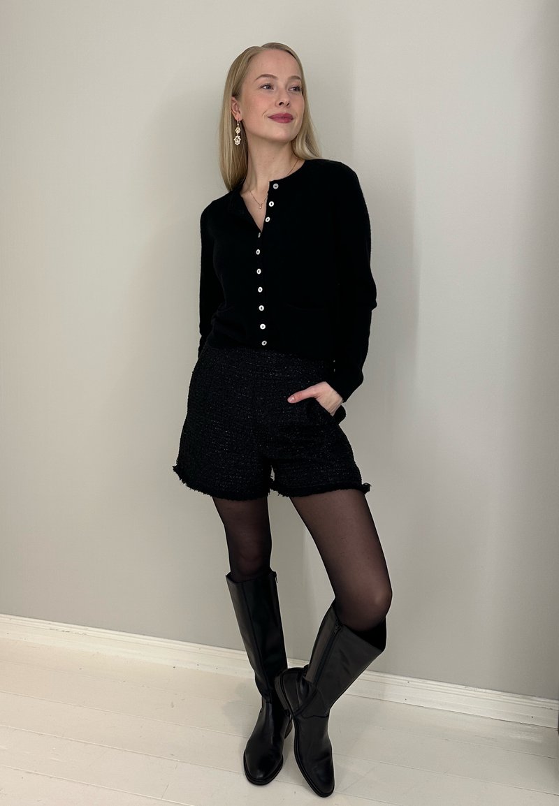 Cardigan noir avec boutons, shorts noirs texturés, collants noirs et bottes en cuir noires montantes jusqu'aux genoux. Design simple et coupe ajustée.
