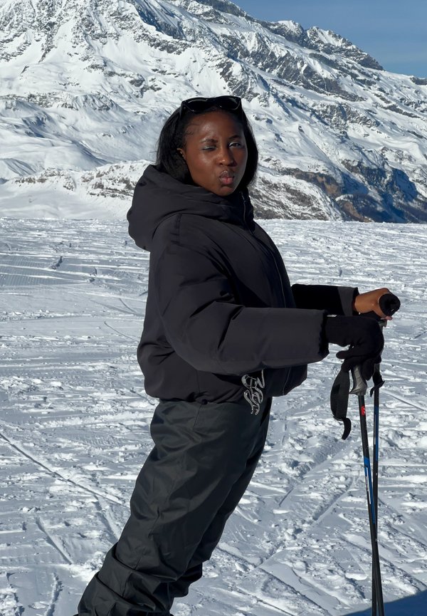 Femme en tenue d'hiver noire tenant des bâtons de ski se tenant sur la neige avec une montagne enneigée en arrière-plan sous un ciel bleu clair.
