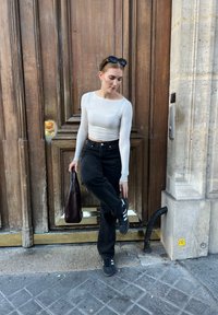 Witte lange mouw top, zwarte wijde jeans en zwarte sneakers met witte strepen. Houdt een donkerbruine tas vast nabij een houten deuropening.