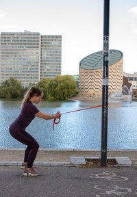 Kobieta w fioletowym stroju sportowym używająca taśmy oporowej przymocowanej do słupa, ćwiczy nad wodą z nowoczesnymi budynkami i drzewami w tle.