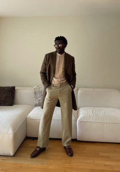 Hombre con abrigo marrón, camisa beige y pantalones color canela, de pie con las manos en los bolsillos frente a un sofá seccional blanco con cojines de estampado animal.