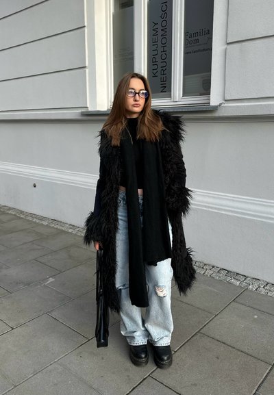 Abrigo de piel sintética negro sobre una blusa negra, jeans ligeros de mezclilla azul desgastados, botas de plataforma negras y una bufanda negra. Unas gafas completan el look.