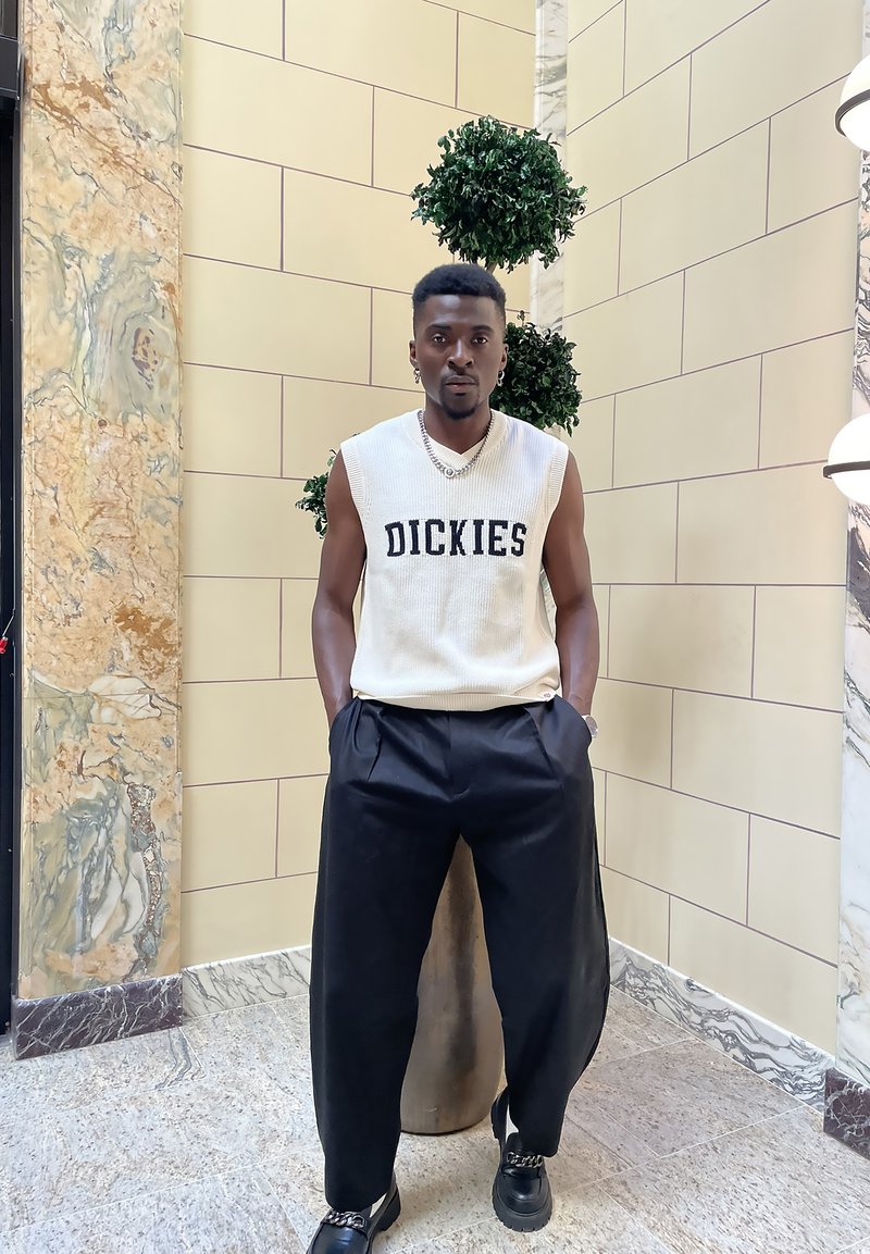Αμάνικο κρεμ πλεκτό τοπ με το λογότυπο "DICKIES", συνδυασμένο με χαλαρούς μαύρους παντελόνους και μαύρα παπούτσια με αλυσίδες. Το φόντο έχει μάρμαρο και πράσινα φυτά.