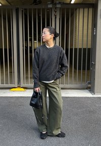 Temno sivi volnen pulover, ohlapne olivno zelene hlače, prozorna očala, večplastni ogrlice, drži črno torbo. Stoji ob ograjenem območju.