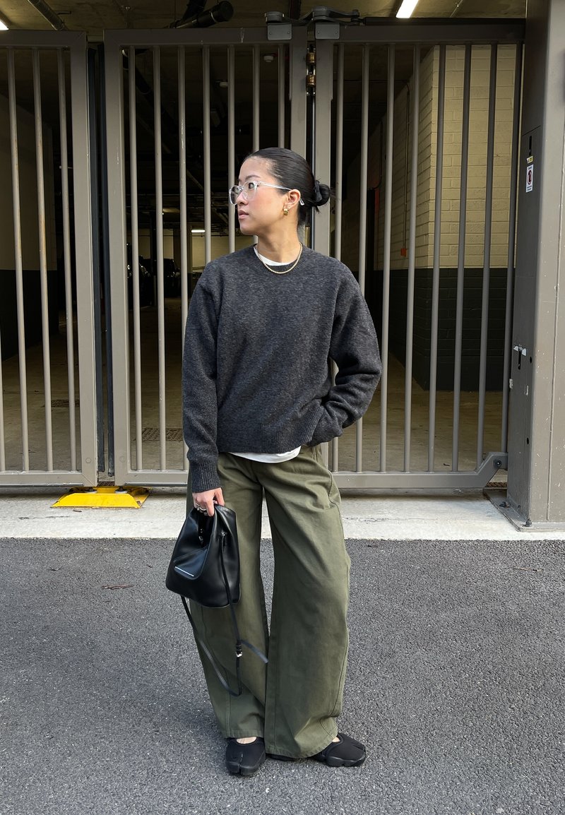 Pull en laine gris foncé, pantalon ample vert olive, lunettes transparentes, colliers superposés, tenant un sac noir. Debout près d'une zone clôturée.