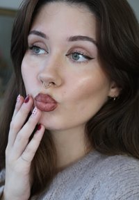 Woman with long brown hair, wearing a gold septum ring, neutral eye makeup, and glossy lips. Clothing is a textured, light fabric.