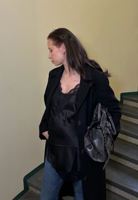 Manteau noir sur un caraco en satin noir orné de dentelle, associé à un jean bleu ; tenant un sac à main sombre en se tenant sur des escaliers.