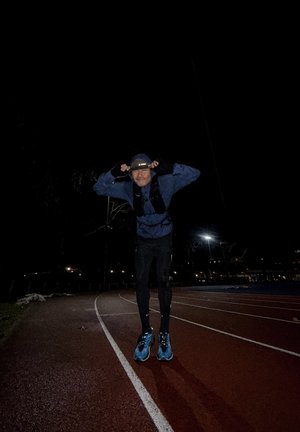 Hombre con chaqueta azul y pantalones negros ajustándose la gorra mientras está de pie en una pista de atletismo al aire libre con poca luz durante la noche.