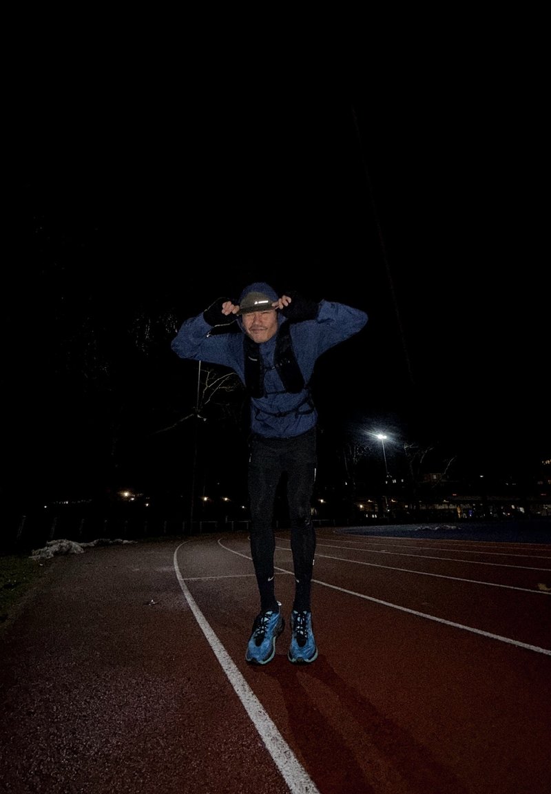 Άνδρας με μπλε μπουφάν και μαύρο παντελόνι, που ρυθμίζει το καπέλο του ενώ στέκεται σε μια σκοτεινά φωτισμένη εξωτερική διαδρομή για τρέξιμο τη νύχτα.