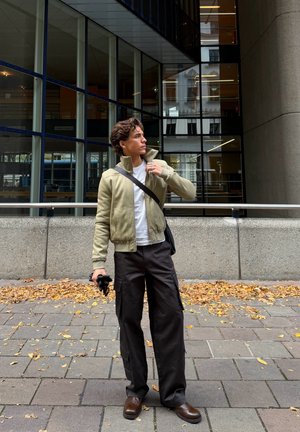 Chaqueta bomber color oliva claro, camiseta blanca, pantalones cargo oscuros, zapatos marrones, sosteniendo una cámara; de pie frente a un edificio moderno con detalles de cristal.