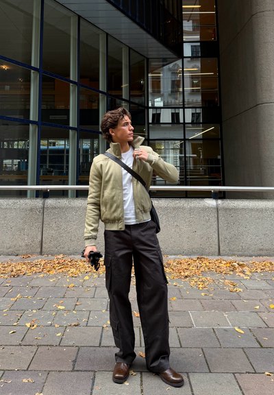 Chaqueta bomber color oliva claro, camiseta blanca, pantalones cargo oscuros, zapatos marrones, sosteniendo una cámara; de pie frente a un edificio moderno con detalles de cristal.