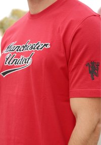 Camiseta de algodón roja con "Manchester United" bordado en blanco en el pecho y un emblema negro en la manga.