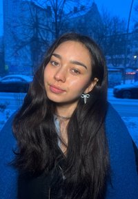 Jeune femme aux longs cheveux noirs et châle bleu, portant des boucles d'oreilles en forme de nœud scintillantes, debout en plein air dans un cadre urbain enneigé en soirée.