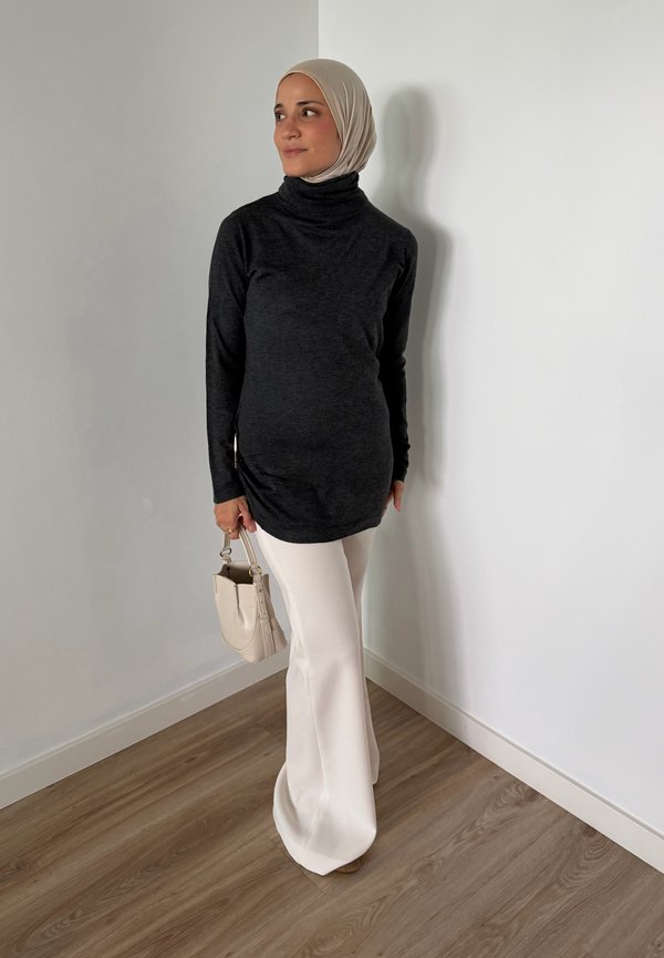 Femme portant un hijab beige, un haut noir à manches longues, un pantalon blanc, tenant un sac à main beige, debout contre un mur blanc sur un sol en bois.