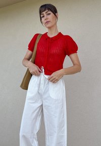 Top roșu, tricotat, cu mânecă scurtă, cu guler și închidere prin nasturi, asortat cu pantaloni albi, largi. O geantă bej este așezată pe un umăr.