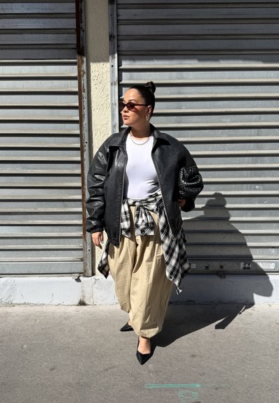 Mujer con gafas de sol lleva una chaqueta de cuero negra, camisa blanca, pantalones beige, camisa de cuadros anudada a la cintura, sosteniendo un clutch negro tejido.