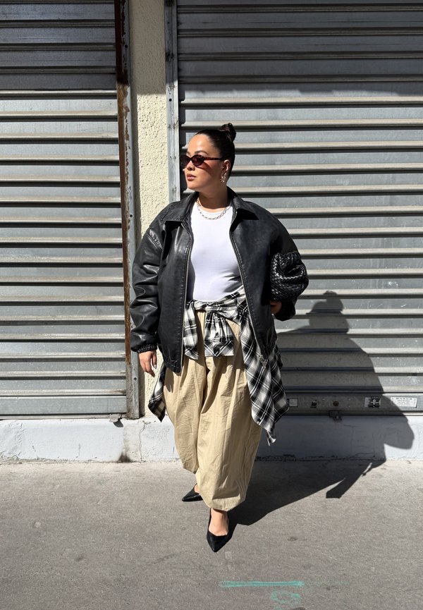 Žena v slnečných okuliaroch nosí čiernu koženú bundu, bielu košeľu, béžové nohavice, kockovanú košeľu zviazanú v páse a drží čiernu pletenú kabelku.