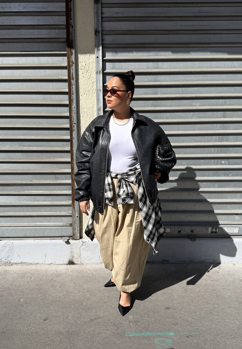 Frau mit Sonnenbrille trägt eine schwarze Lederjacke, ein weisses Hemd, beige Hosen, ein kariertes Hemd um die Taille gebunden und hält eine schwarze gewebte Clutch.