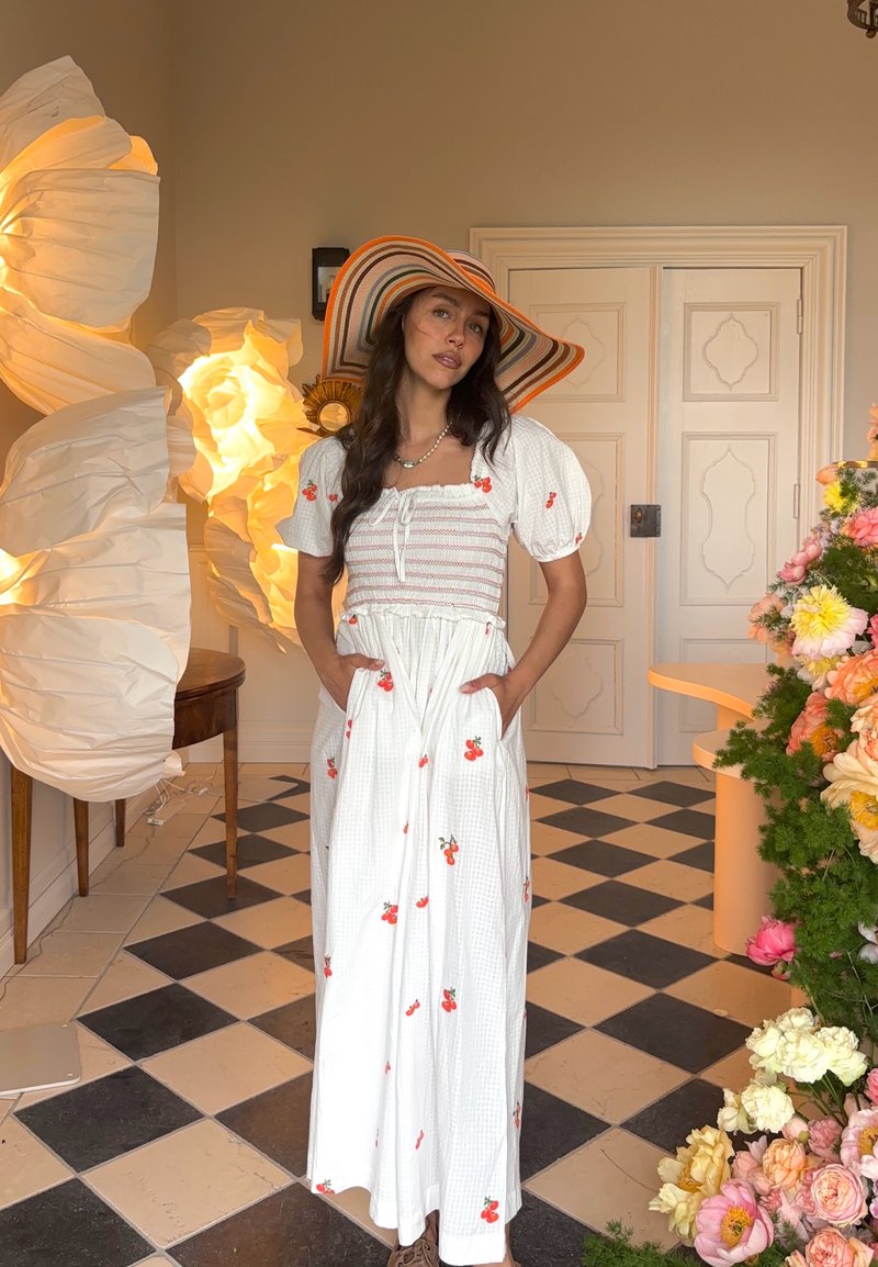 Femme en robe blanche à motifs floraux rouges et chapeau rayé à large bord, debout dans une pièce décorée avec des lampes en papier et des fleurs.