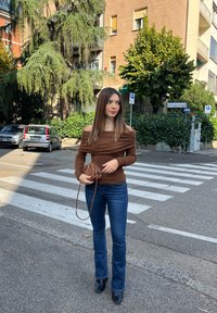 Brown off-the-shoulder top with draped fabric, blue flared jeans, black ankle boots, and a brown handbag, on a city street with trees.