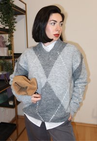 Pull en mélange de laine à motif losanges gris et gris clair, avec col en V et poignets côtelés. Tenant un sac en daim marron.