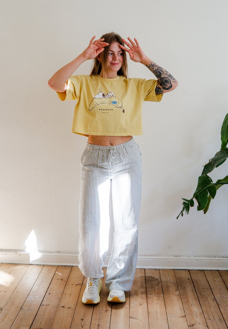 Jeune femme au bras tatoué portant un crop top jaune Peanuts et un pantalon à rayures, posant à l'intérieur près d'une plante verte sur un sol en bois.