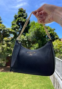 Main tenant un sac à main en cuir noir Michael Kors avec une bandoulière en chaîne dorée devant un fond d'arbres verts et de ciel bleu en plein air.