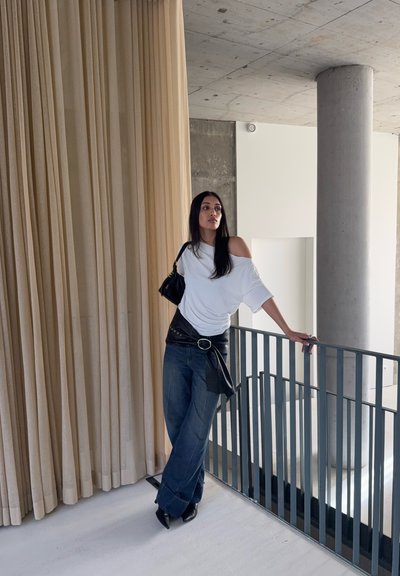 Mujer con cabello largo y oscuro, vestida con blusa blanca fuera del hombro, jeans anchos, botas negras y cinturón negro en la cintura, apoyada en una barandilla en un espacio interior moderno.