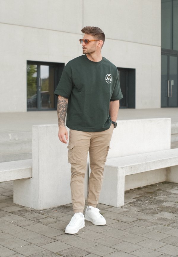Muž s tetovanou rukou, který má na sobě sluneční brýle, tmavě zelené oversized tričko, béžové cargo kalhoty a bílé tenisky, stojící u betonových laviček venku.