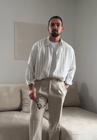 Homme portant une chemise blanche à rayures et un pantalon beige se tient à l'intérieur, tenant des lunettes noires à côté de lui près d'un canapé beige.