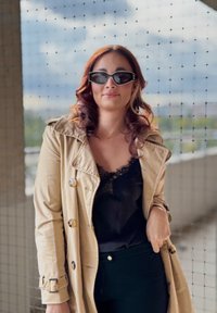 Beige Trenchcoat mit silbernen Knöpfen über einem schwarzen Spitzenoberteil, kombiniert mit schwarzen Hosen und kantigen dunklen Sonnenbrillen. Hintergrund aus Netz.