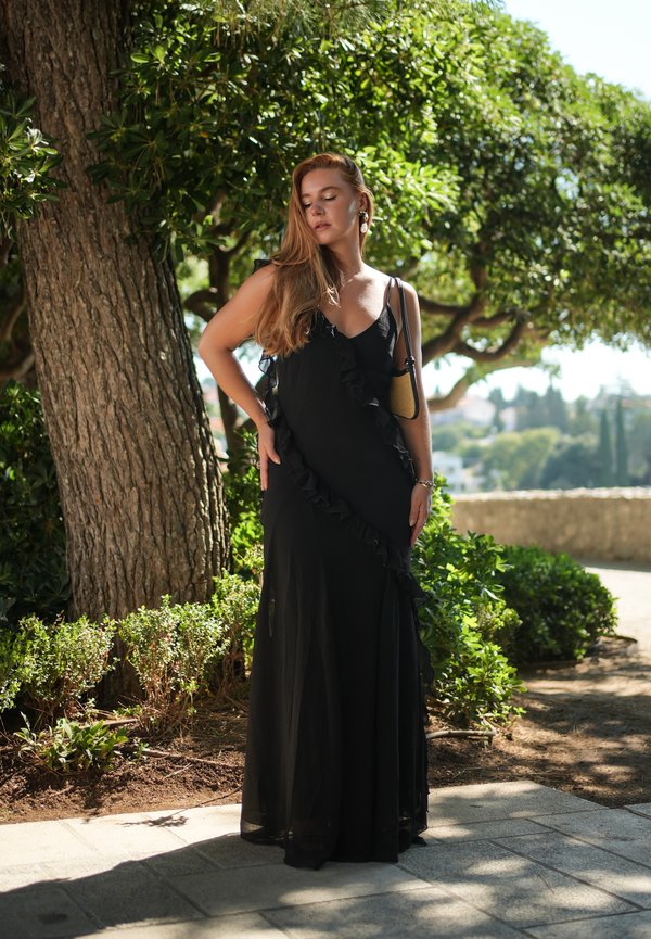 Donna con i capelli lunghi che indossa un vestito nero senza maniche con balze e una borsa a tracolla, in piedi vicino a un albero con verdura sullo sfondo.