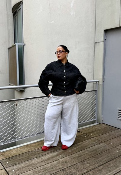 Mujer de pie en una terraza de madera con una chaqueta negra abotonada, pantalones anchos blancos, zapatos rojos, gafas y sosteniendo un bolso negro con textura.