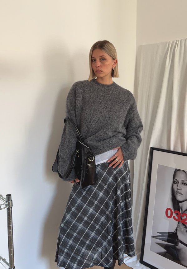 Femme en pull gris pelucheux et jupe à carreaux debout à l'intérieur, tenant un sac en cuir noir, avec un portrait en noir et blanc encadré appuyé à proximité.