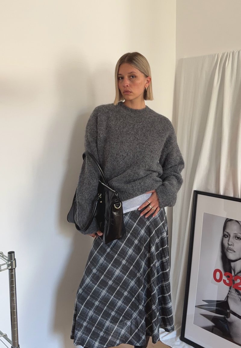 Woman in a gray fuzzy sweater and plaid skirt stands indoors holding a black leather bag, with framed black-and-white portrait leaning nearby.