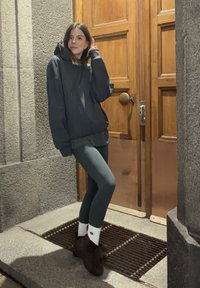 Prevelik temen pulover s kapuco čez siv zgornji del, kombiniran z rebrastimi zelenimi legicami in rjavimi škornji, stoji ob lesenih vratih.