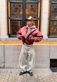 Raudonas satino bombardieriaus striukė vilkima ant pilko džemperio su kapišonu, derinama su laisvomis pilkomis sportinėmis kelnėmis ir tamsiais batais, stovint prie didelių langų.