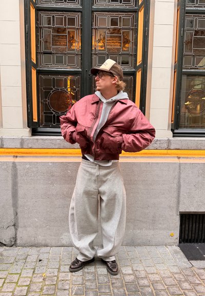 Chaqueta bomber de satén roja sobre una sudadera con capucha gris, combinada con pantalones deportivos grises holgados y zapatos oscuros, de pie frente a grandes ventanas.