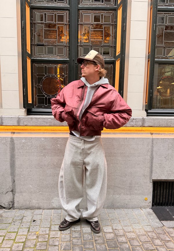 Raudonas satino bombardieriaus striukė vilkima ant pilko džemperio su kapišonu, derinama su laisvomis pilkomis sportinėmis kelnėmis ir tamsiais batais, stovint prie didelių langų.