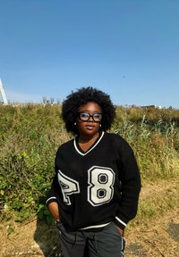 Melnā adīta džemperis ar baltām svītrām un numuriem "8" un "P" priekšpusē, savienots ar pelēkiem biksēm. Dabisks fons aizmugurē.