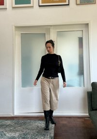 Crna bluzica dugih rukava, bež široke pamučne hlače, crne čizme do koljena i torba preko ramena; svijetao unutarnji prostor s staklenim vratima.