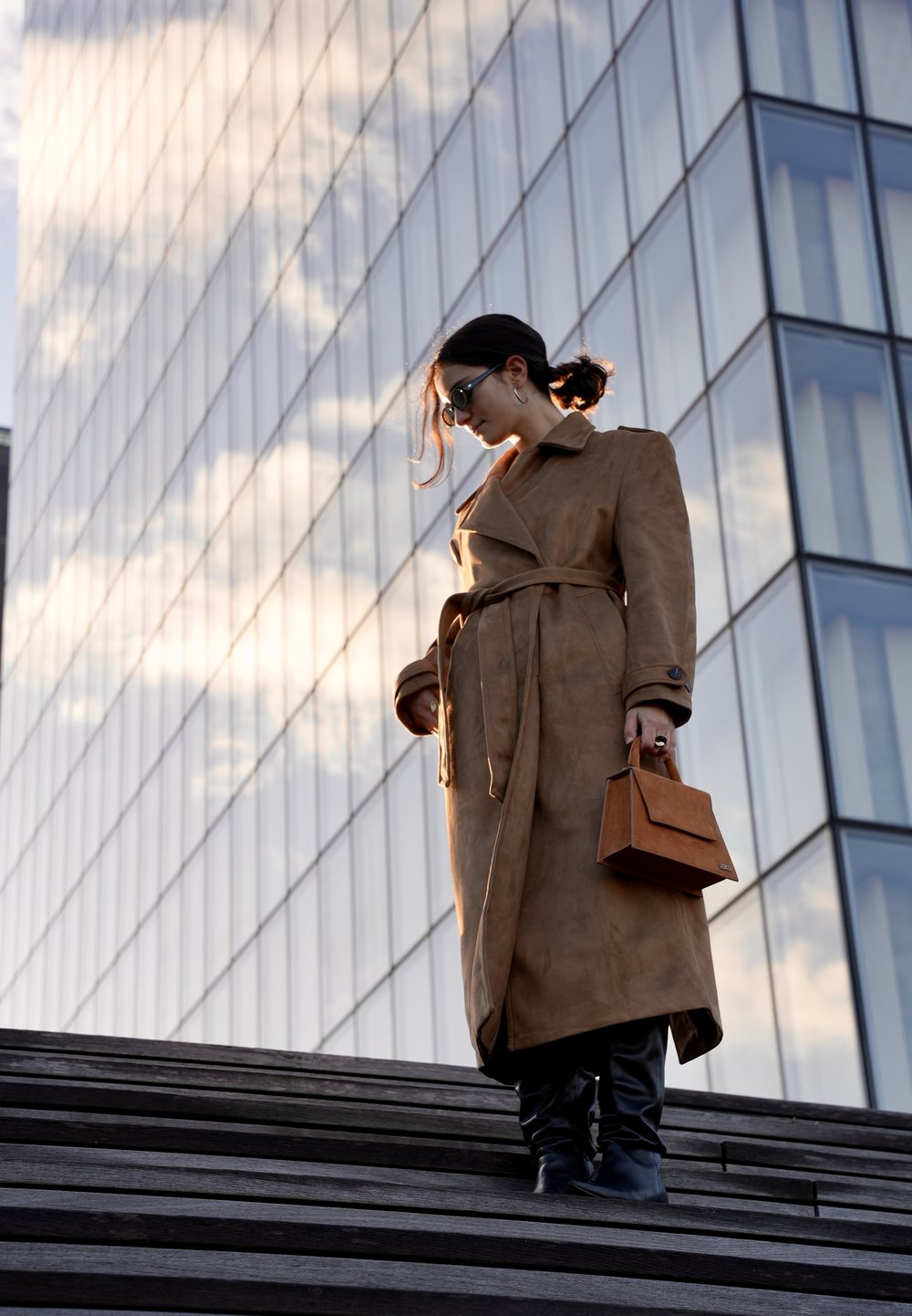 Beige suède trenchcoat met een ceintuur in de taille, gecombineerd met zwarte leren broek en bijpassende laarzen. Houdt een kleine bruine handtas vast, tegen een glazen achtergrond.