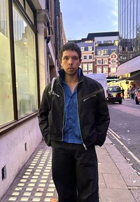 Must bomberjakk hõbedaste tõmblukkude detailidega, kantud sinise särgi peal. Mustad püksid, vabaaja lõikega, seisab linna tänaval hämarikus.