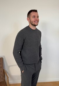 Homme souriant regardant vers la droite, portant un pull et un pantalon gris foncé, debout à l'intérieur à côté d'une chaise en bois contre un mur uni.