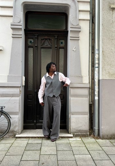 Chaleco gris sobre una camisa rosa clara, pantalones grises anchos y zapatos oscuros. De pie junto a una puerta marrón con paneles de cristal, al lado de una bicicleta.