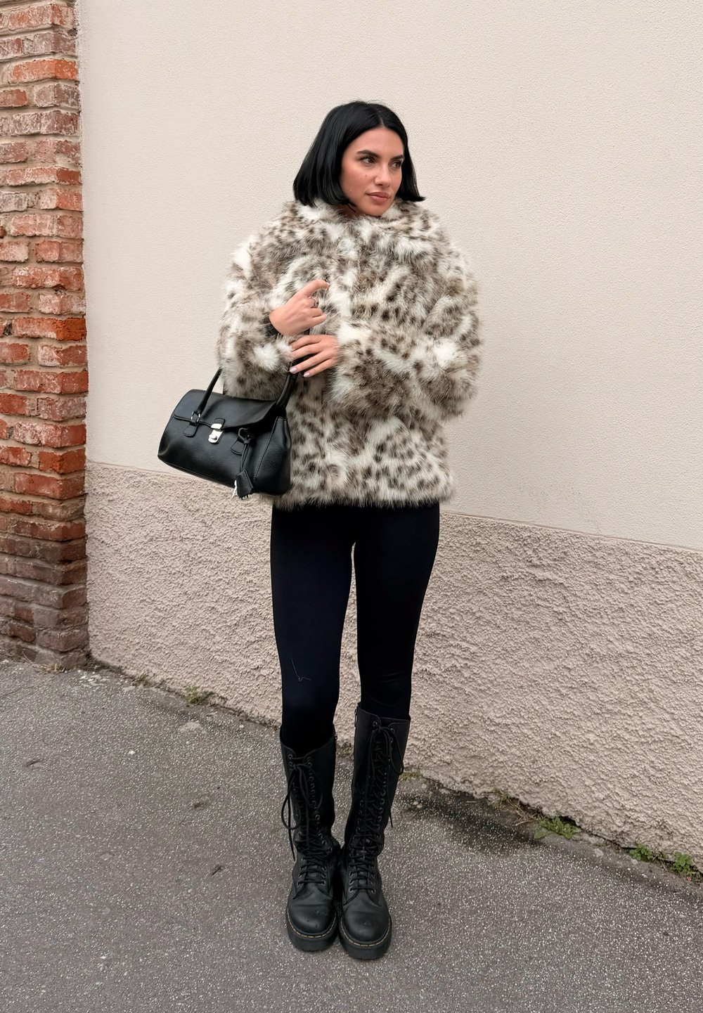 Faux fur jacket in light tan with dark spots, paired with black leggings and knee-high lace-up boots. Black handbag with silver closure.